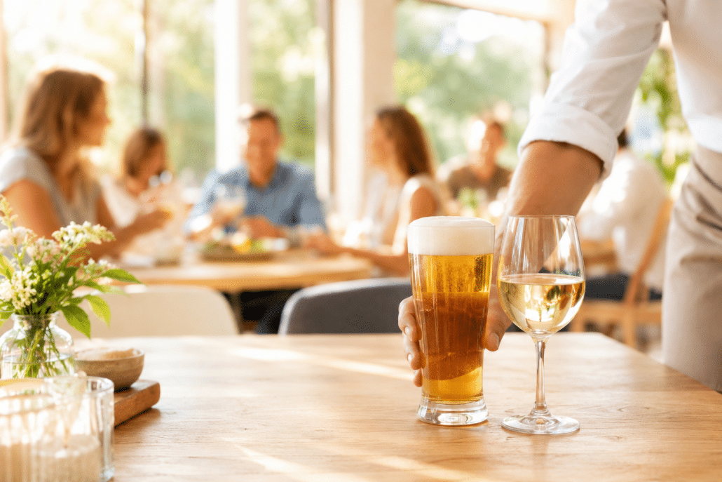 Eine Servicekraft stellt ein frisch gezapftes Bier und ein Glas Weißwein auf einem hellen Holztisch in einem freundlichen Restaurant ab. Im Hintergrund sitzen unscharf Gäste in entspannter Atmosphäre bei warmem Tageslicht.