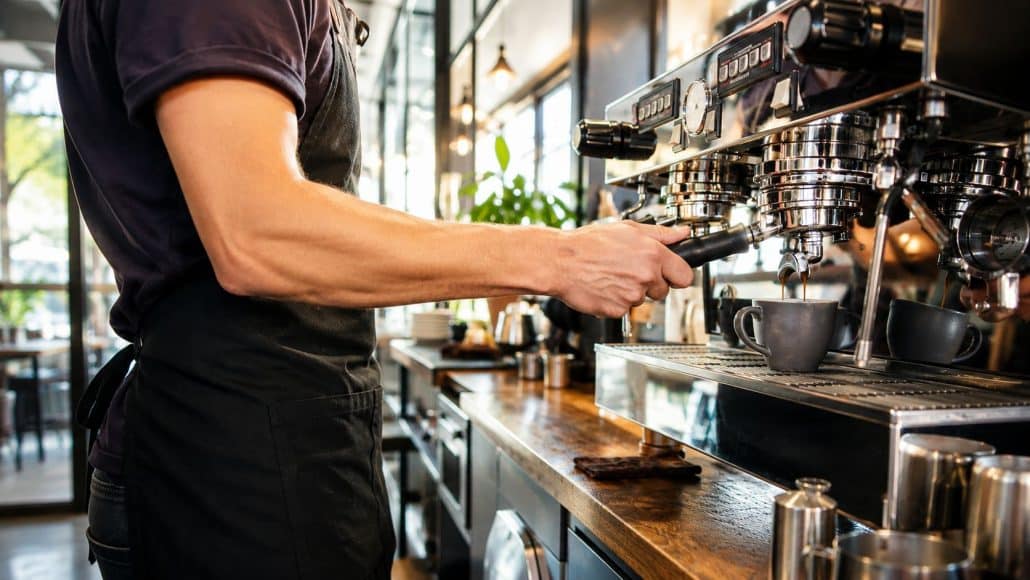 Ein Barista bereitet an einer professionellen Siebträgermaschine frischen Espresso zu. Das moderne Café wird von warmem Tageslicht durch große Fenster erhellt und vermittelt eine einladende, hochwertige Atmosphäre. Die glänzende Kaffeemaschine, natürliche Materialien und die offene Raumgestaltung unterstreichen die Professionalität und den modernen Charakter des Betriebs.