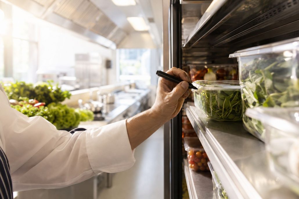 Koch beschriftet frische Lebensmittel und räumt sie nach dem FIFO-Prinzip in ein Kühlregal einer professionellen Restaurantküche ein.